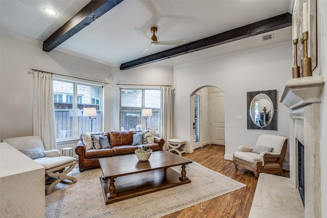 living room featuring wood-type flooring, beam ceiling, a fireplace, ornamental molding, and ceiling fan