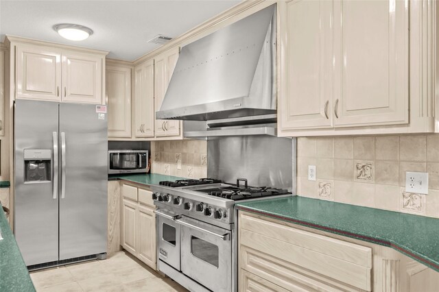 kitchen with stainless steel appliances, wall chimney range hood, backsplash, cream cabinetry, and light tile patterned floors