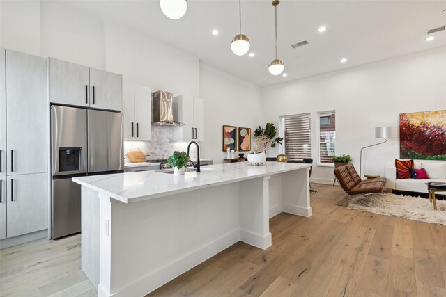 kitchen featuring light wood-type flooring, stainless steel fridge, a center island with sink, and wall chimney range hood