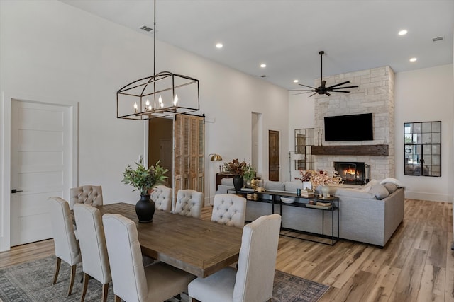 dining room featuring light hardwood / wood-style floors, a stone fireplace, a high ceiling, and ceiling fan with notable chandelier