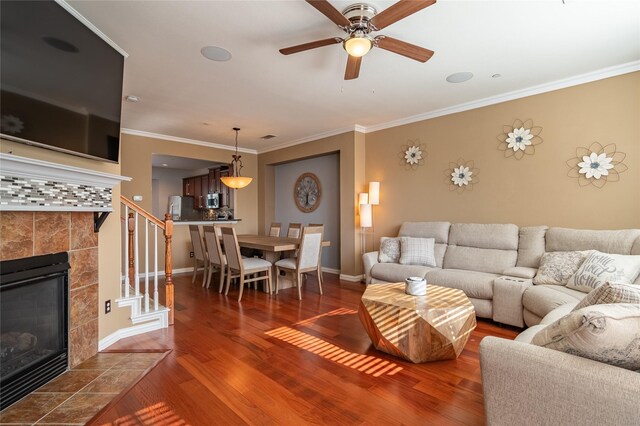 living room featuring a tiled fireplace, dark wood-type flooring, ornamental molding, and ceiling fan