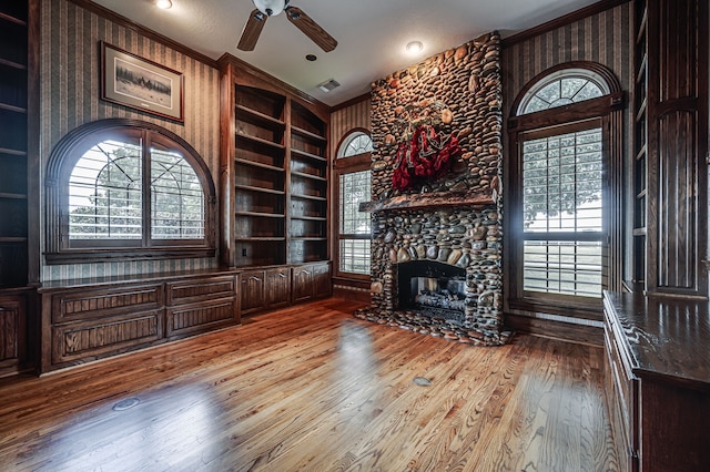 unfurnished living room with light wood-type flooring, a stone fireplace, built in features, ceiling fan, and ornamental molding