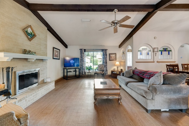 living room featuring a brick fireplace, ceiling fan, beamed ceiling, and light hardwood / wood-style flooring