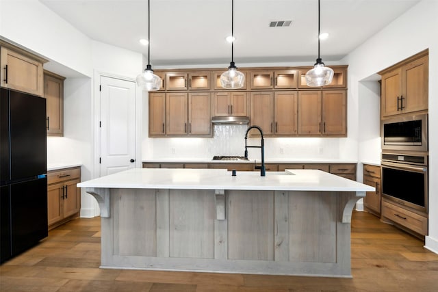 kitchen featuring decorative backsplash, appliances with stainless steel finishes, a kitchen island with sink, and wood-type flooring