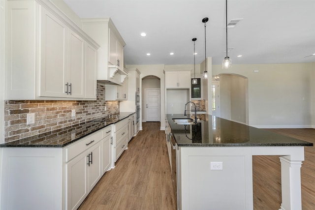 kitchen with pendant lighting, an island with sink, sink, dark stone countertops, and light wood-type flooring