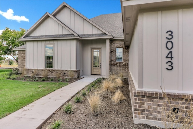 doorway to property with brick siding, a yard, board and batten siding, and roof with shingles