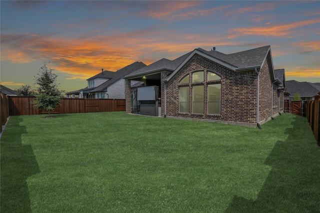 back of property at dusk with a yard, brick siding, roof with shingles, and a fenced backyard