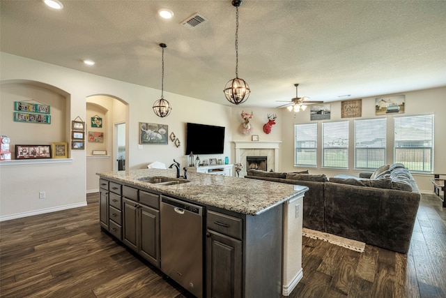 kitchen featuring dishwasher, a tiled fireplace, ceiling fan with notable chandelier, dark hardwood / wood-style flooring, and a kitchen island with sink