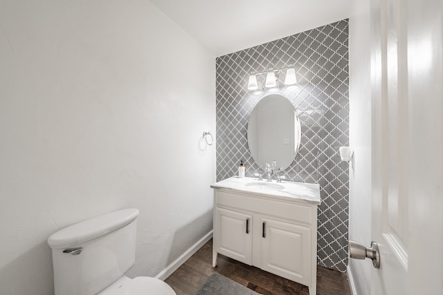 half bathroom with toilet, wood finished floors, vanity, baseboards, and wallpapered walls