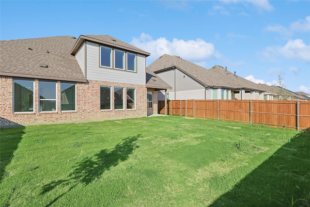 rear view of property featuring roof with shingles, brick siding, a lawn, and a fenced backyard