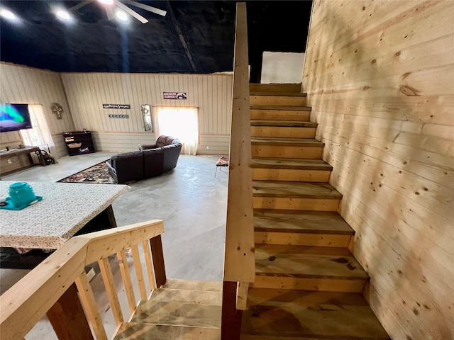 stairway featuring unfinished concrete floors, wood walls, and a ceiling fan