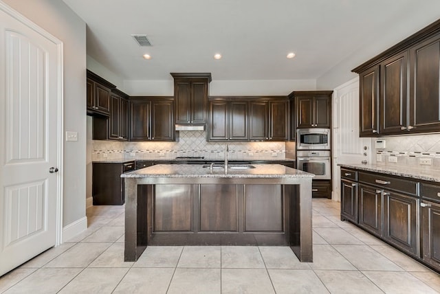 kitchen with an island with sink, stainless steel appliances, light stone counters, and decorative backsplash