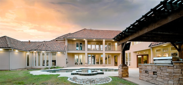 back house at dusk featuring a balcony, a patio area, an outdoor kitchen, a lawn, and french doors