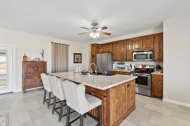 kitchen featuring appliances with stainless steel finishes, light stone counters, an island with sink, ceiling fan, and a kitchen bar