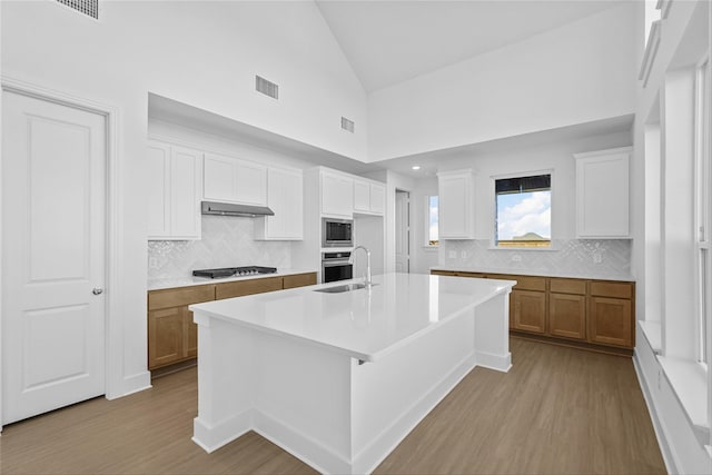 kitchen featuring a kitchen island with sink, high vaulted ceiling, white cabinets, ventilation hood, and appliances with stainless steel finishes