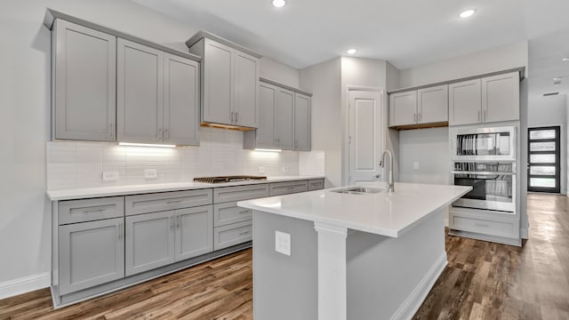 kitchen with wood-type flooring, sink, decorative backsplash, appliances with stainless steel finishes, and a center island with sink