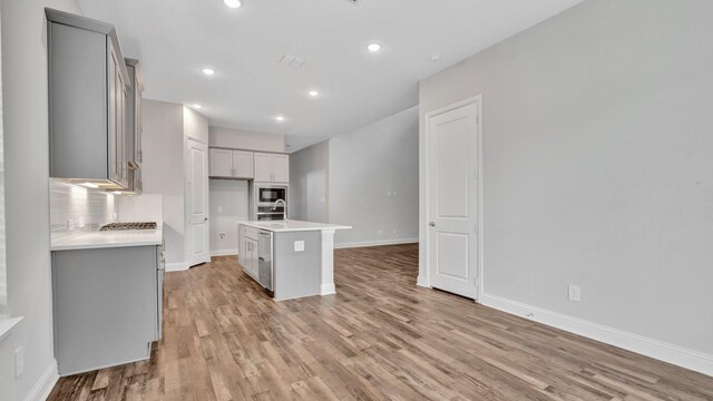 kitchen featuring light wood-type flooring, gray cabinets, an island with sink, decorative backsplash, and black microwave