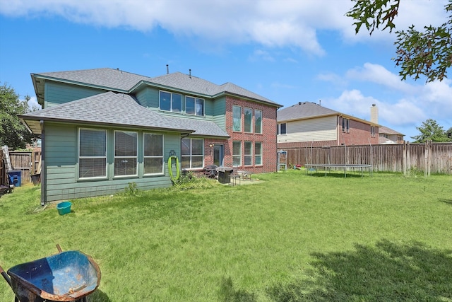 back of house with a trampoline, roof with shingles, brick siding, a lawn, and fence