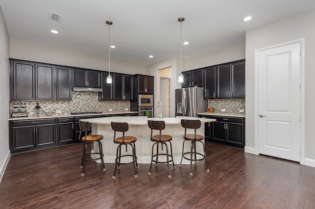 kitchen featuring appliances with stainless steel finishes, backsplash, hanging light fixtures, an island with sink, and dark hardwood / wood-style flooring