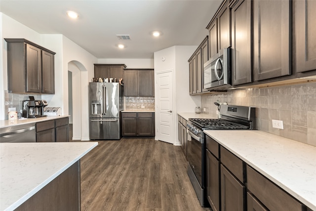 kitchen featuring light stone counters, stainless steel appliances, dark brown cabinets, dark wood-type flooring, and decorative backsplash