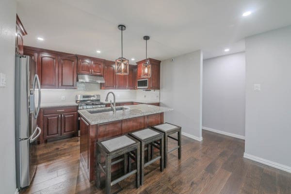 kitchen with stainless steel appliances, decorative light fixtures, a center island with sink, and dark hardwood / wood-style flooring