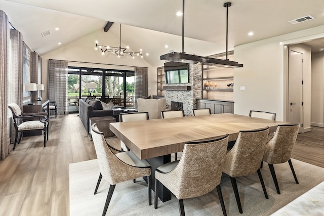 dining area featuring beam ceiling, an inviting chandelier, a stone fireplace, high vaulted ceiling, and light hardwood / wood-style floors