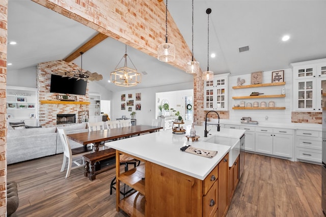 kitchen with white cabinets, decorative light fixtures, vaulted ceiling with beams, a kitchen island with sink, and a brick fireplace