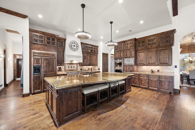 kitchen with built in appliances, backsplash, a center island with sink, and wood-type flooring