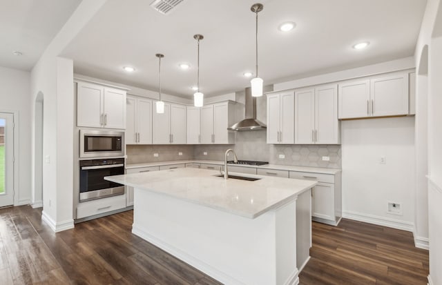 kitchen featuring wall chimney exhaust hood, dark hardwood / wood-style floors, an island with sink, light stone counters, and stainless steel appliances