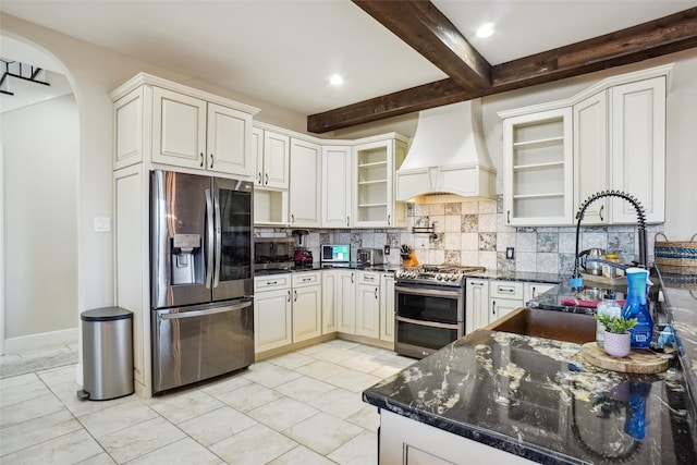 kitchen featuring beamed ceiling, custom range hood, stainless steel appliances, decorative backsplash, and dark stone countertops