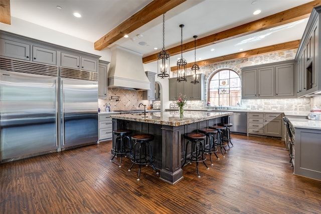 kitchen featuring a kitchen bar, light stone counters, appliances with stainless steel finishes, custom range hood, and a center island with sink