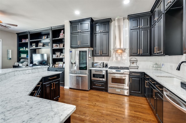 kitchen with light stone countertops, wall chimney exhaust hood, appliances with stainless steel finishes, light hardwood / wood-style floors, and ceiling fan