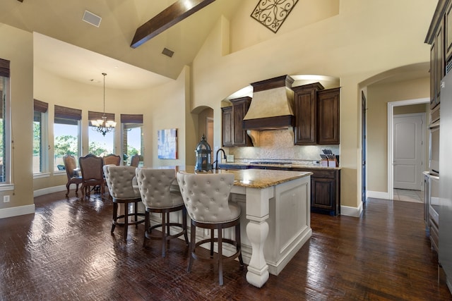 kitchen with dark hardwood / wood-style flooring, decorative light fixtures, custom range hood, high vaulted ceiling, and a kitchen island with sink