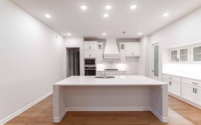 kitchen with stainless steel oven, custom range hood, built in microwave, a center island with sink, and white cabinets