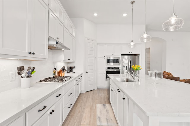 kitchen with decorative light fixtures, appliances with stainless steel finishes, a kitchen island with sink, sink, and light wood-type flooring