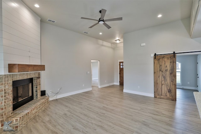 unfurnished living room featuring a brick fireplace, a barn door, light hardwood / wood-style floors, ceiling fan, and a towering ceiling