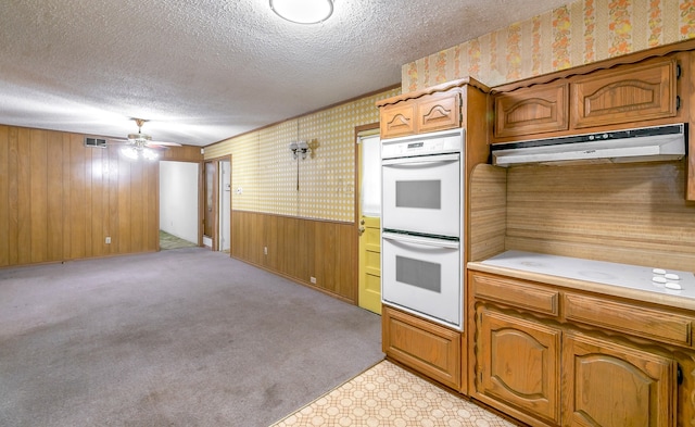 kitchen featuring wooden walls, light colored carpet, double oven, ceiling fan, and a textured ceiling