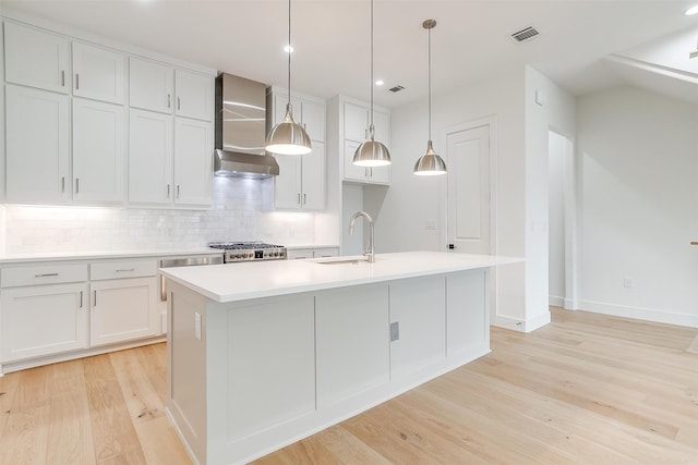 kitchen featuring decorative light fixtures, wall chimney exhaust hood, an island with sink, sink, and light wood-type flooring