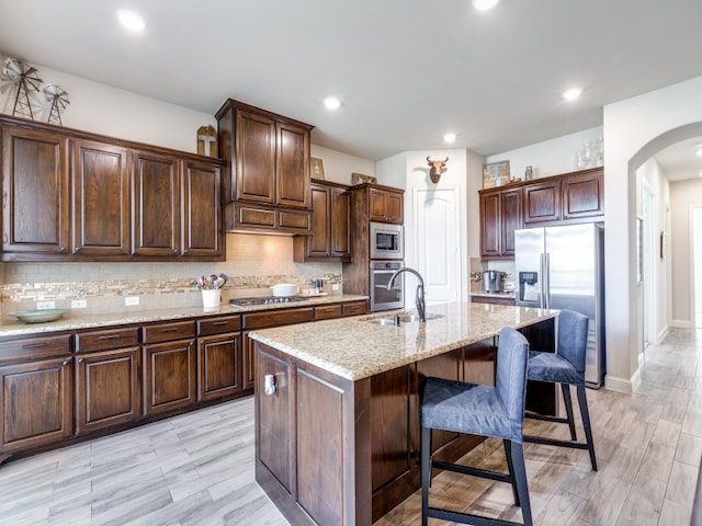 kitchen with a center island with sink, dark brown cabinetry, appliances with stainless steel finishes, light stone counters, and tasteful backsplash