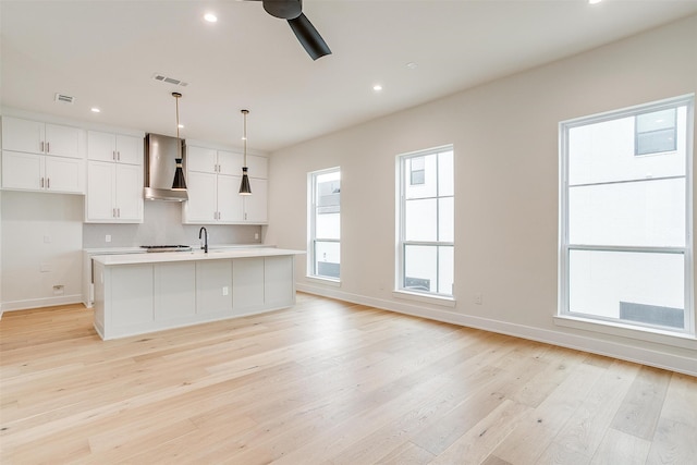 kitchen with a kitchen island with sink, white cabinetry, ceiling fan, wall chimney range hood, and light wood-type flooring