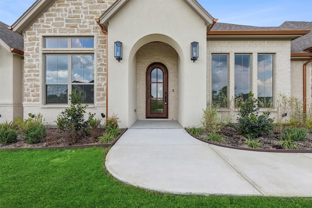 doorway to property featuring stone siding, a yard, roof with shingles, and stucco siding