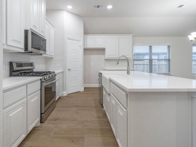 kitchen featuring appliances with stainless steel finishes, tasteful backsplash, a center island with sink, light hardwood / wood-style floors, and white cabinetry