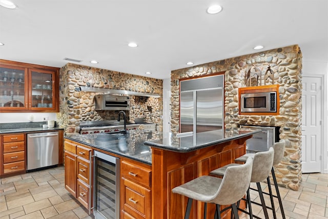 kitchen with built in appliances, a breakfast bar area, a center island with sink, beverage cooler, and dark stone countertops