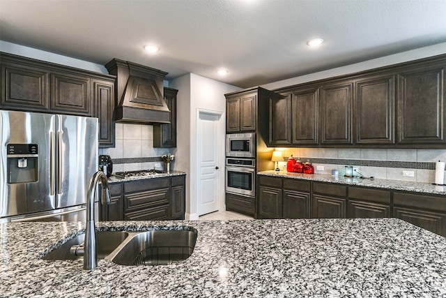 kitchen featuring stone countertops, backsplash, premium range hood, sink, and appliances with stainless steel finishes