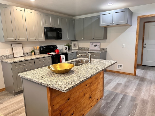 kitchen featuring light wood-type flooring, sink, a kitchen island with sink, black appliances, and light stone countertops