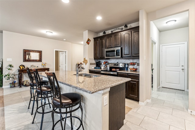 kitchen featuring an island with sink, sink, light stone counters, stainless steel appliances, and dark brown cabinets