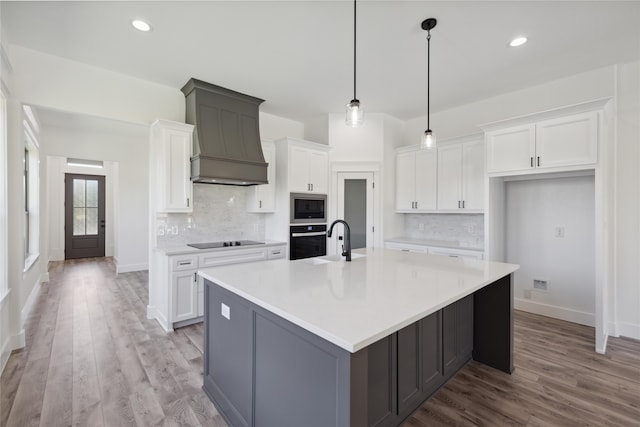 kitchen featuring wall oven, premium range hood, an island with sink, black electric cooktop, and white cabinets