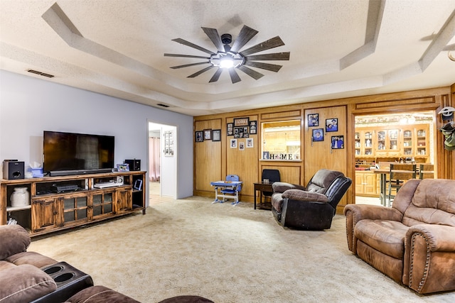 carpeted living room featuring ceiling fan, a textured ceiling, a tray ceiling, and wood walls