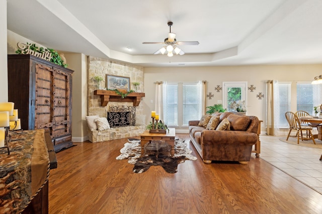 living room featuring a tray ceiling, wood-type flooring, ceiling fan, and a stone fireplace