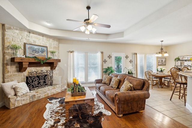 living room with ceiling fan with notable chandelier, a raised ceiling, light hardwood / wood-style flooring, and a stone fireplace